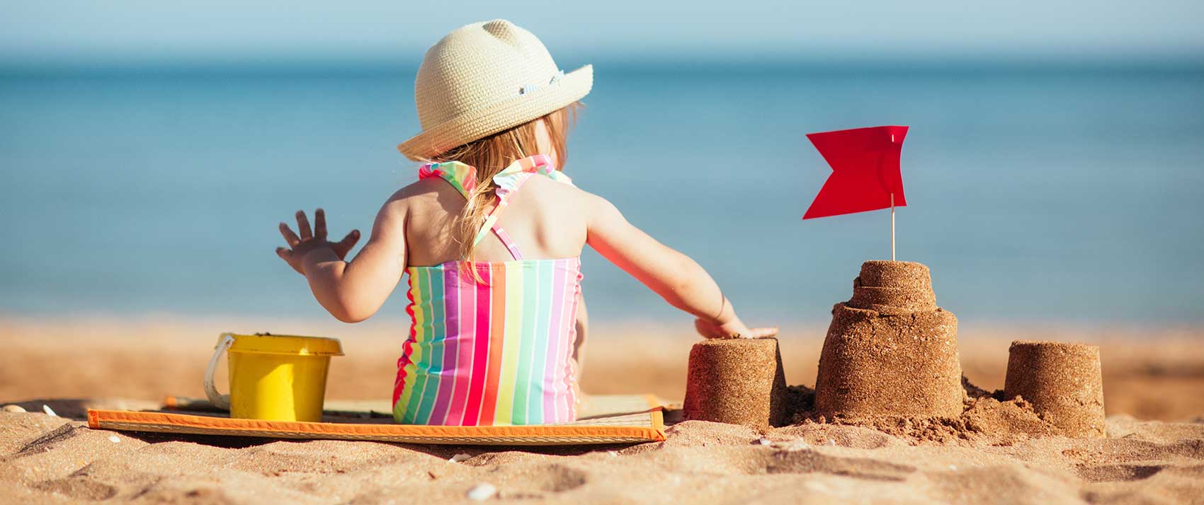  toddler girls sits on beach next to sand castle and bucket